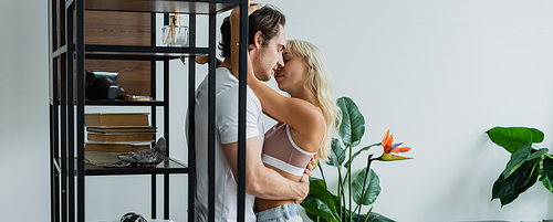 blonde woman hugging man with closed eyes near rack in living room, banner