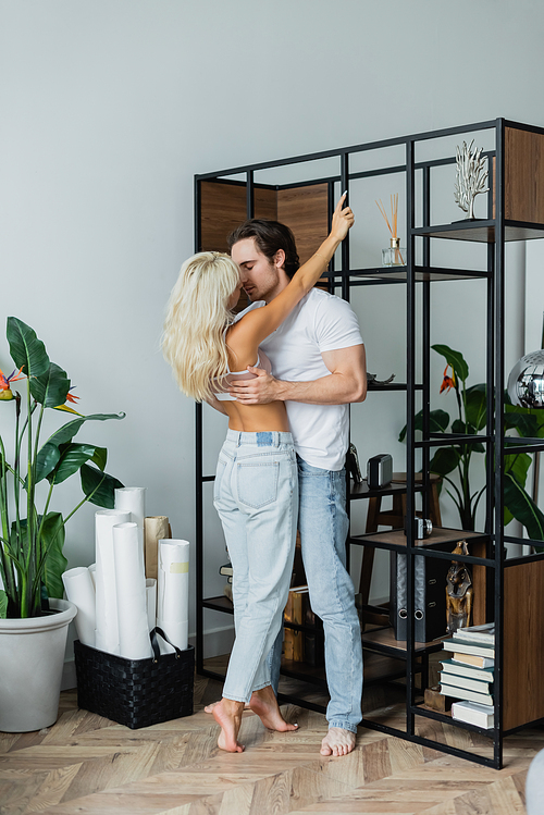 full length of blonde woman hugging man with closed eyes near rack in living room
