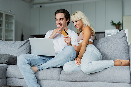 cheerful man holding credit card near girlfriend while looking at laptop and doing online shopping