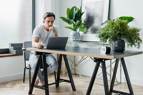 concentrated freelancer looking at laptop while holding smartphone and working from home