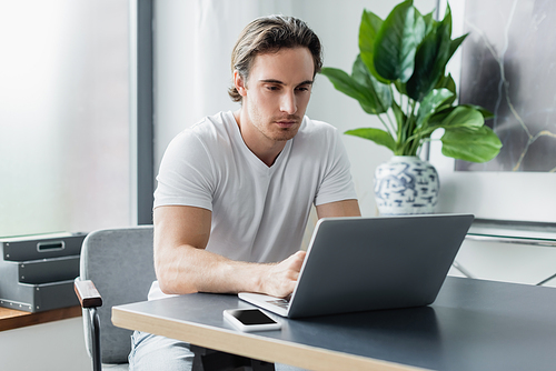 concentrated freelancer looking at laptop near smartphone on desk while working from home