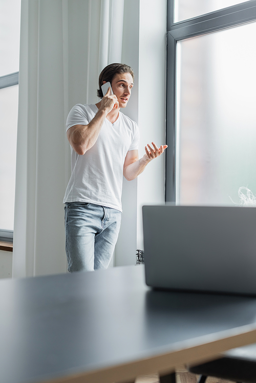 young man gesturing while talking on smartphone near blurred laptop on desk
