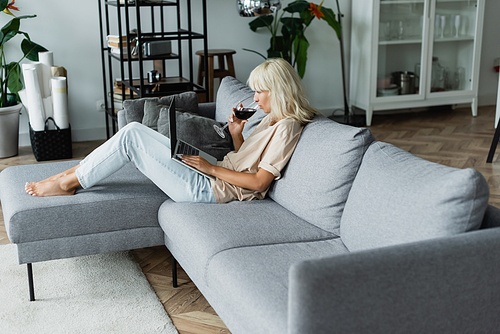 blonde woman drinking red wine while using laptop in living room