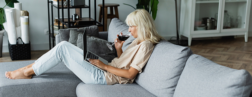 blonde woman drinking red wine while using laptop in living room, banner