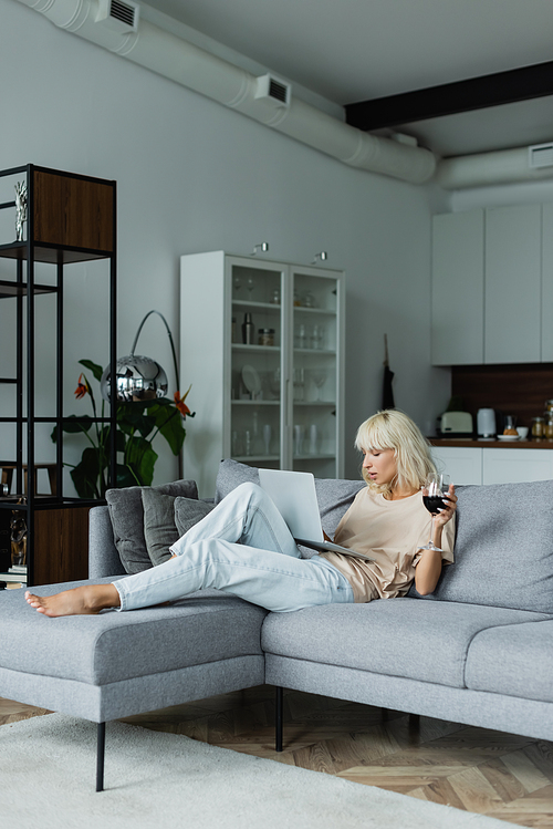 blonde woman holding glass of red wine while using laptop in living room