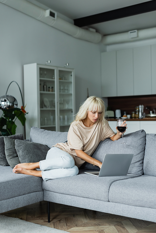 barefoot at blonde woman holding glass of red wine while using laptop in living room