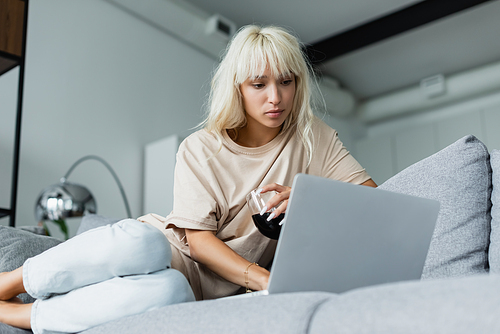 blonde freelancer holding glass of red wine while using laptop in living room