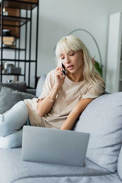 blonde freelancer talking on smartphone near blurred laptop on couch