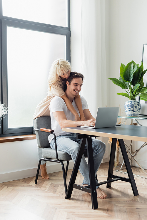 blonde woman hugging happy boyfriend using laptop while working from home