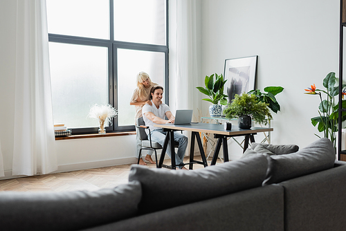 blonde woman massaging shoulders of boyfriend working from home with laptop