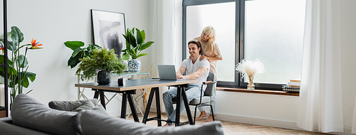 blonde woman massaging shoulders of boyfriend working from home with laptop, banner