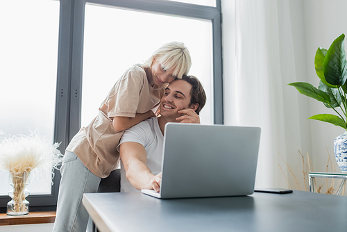 blonde woman hugging smiling boyfriend using laptop while working from home