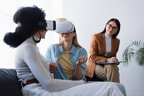 happy interracial lesbian couple in vr headsets holding hands near blurred psychologist writing on clipboard