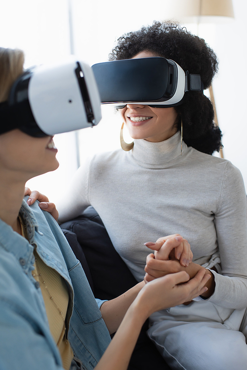 young and happy multiethnic lesbian women gaming in vr headsets while holding hands at home