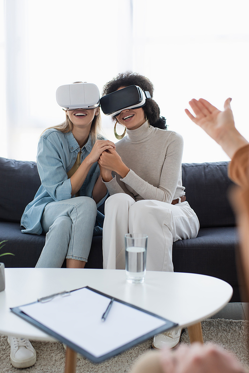 smiling multiethnic lesbian couple in vr headsets holding hands near blurred psychologist pointing with hand