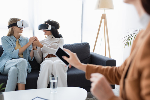smiling same sex couple in vr headsets holding hands on sofa near blurred psychologist with smartphone