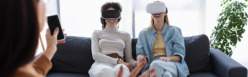 interracial lesbian couple in vr headsets smiling near blurred psychologist with smartphone, banner