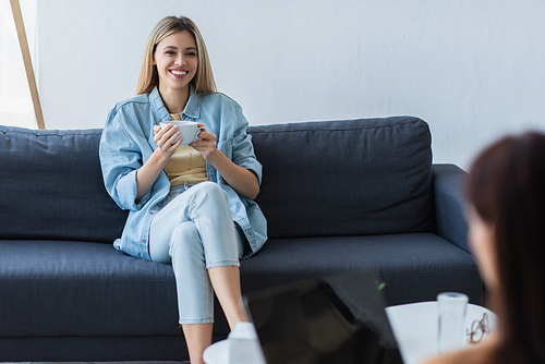 smiling woman sitting on couch with cup of tea during consultation with blurred psychologist