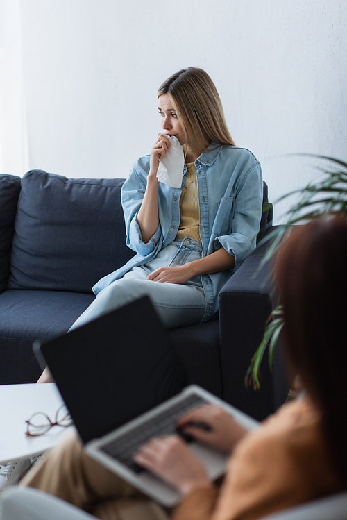 depressed woman sitting on couch near psychologist with laptop on blurred foreground
