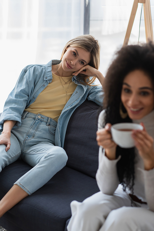 happy lesbian woman looking at blurred african american girlfriend drinking tea on couch