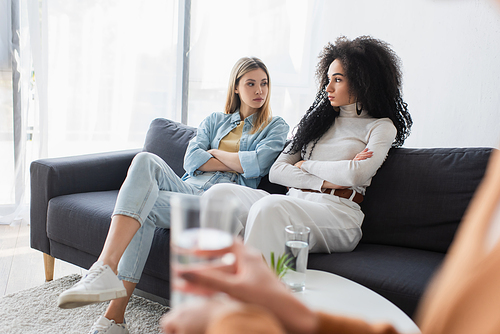 offended interracial lesbians sitting with crossed arms on couch during psychological consultation