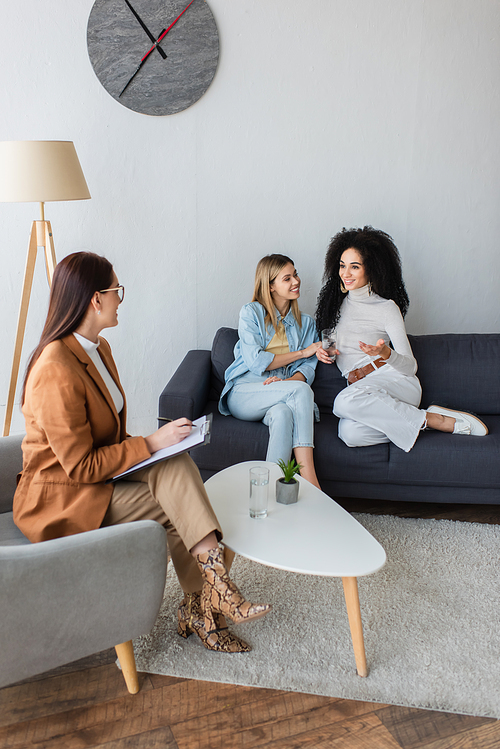 psychologist with clipboard listening to smiling interracial lesbian couple sitting on couch in consulting room