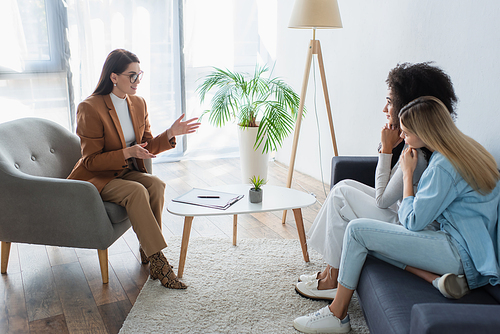 positive psychologist gesturing while talking to interracial couple of lesbians in consulting room