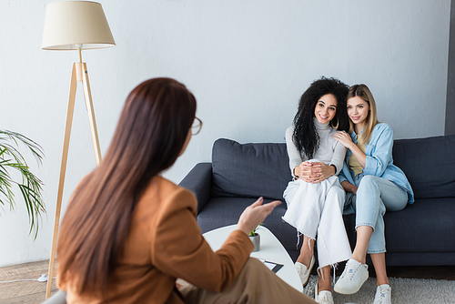 blurred psychologist pointing with hand while talking to happy multiethnic lesbians sitting on couch