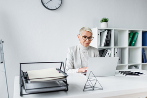 businesswoman with grey hair in glasses and suit using laptop in office