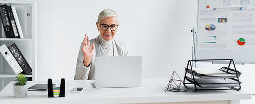 happy senior businesswoman in glasses waving hand during video call on laptop, banner