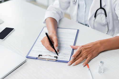 cropped view of doctor writing prescription near gadgets on desk