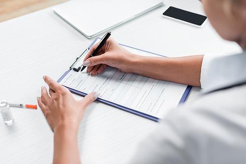 cropped view of blurred doctor writing prescription near gadgets on desk