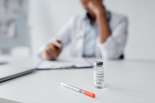 bottle with coronavirus vaccine and syringe on desk near blurred doctor on background