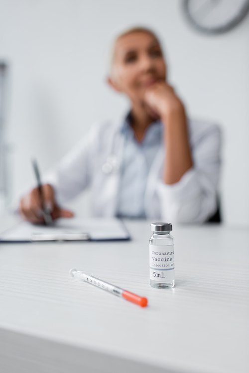 bottle with coronavirus vaccine near syringe and blurred doctor on background