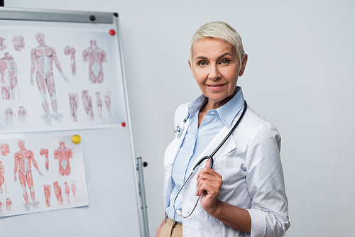 happy senior doctor in white coat with stethoscope standing near flip chart with anatomical pictures