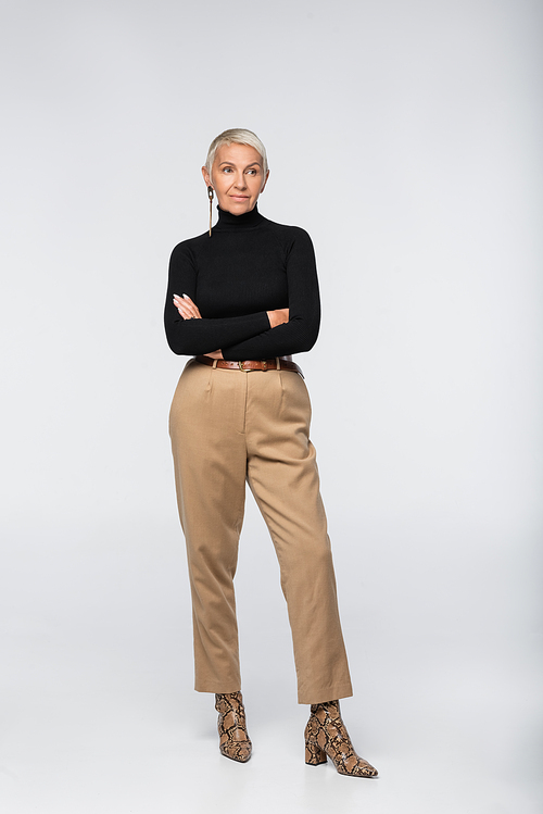 full length of trendy senior woman in beige pants, black turtleneck and animal  boots posing with crossed arms on grey