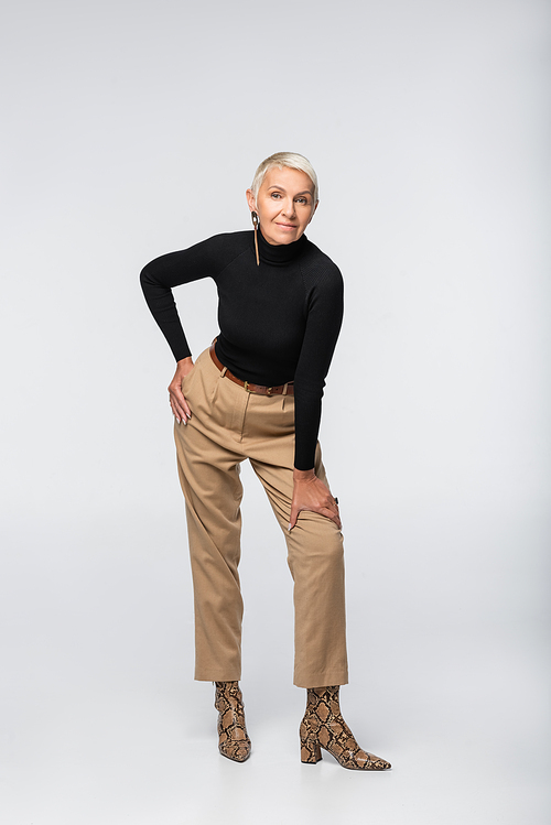 full length of pleased and trendy senior woman in beige pants, black turtleneck and animal print boots posing on grey