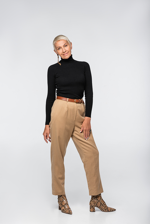 full length of smiling y senior woman in beige pants, black turtleneck and animal  boots posing on grey