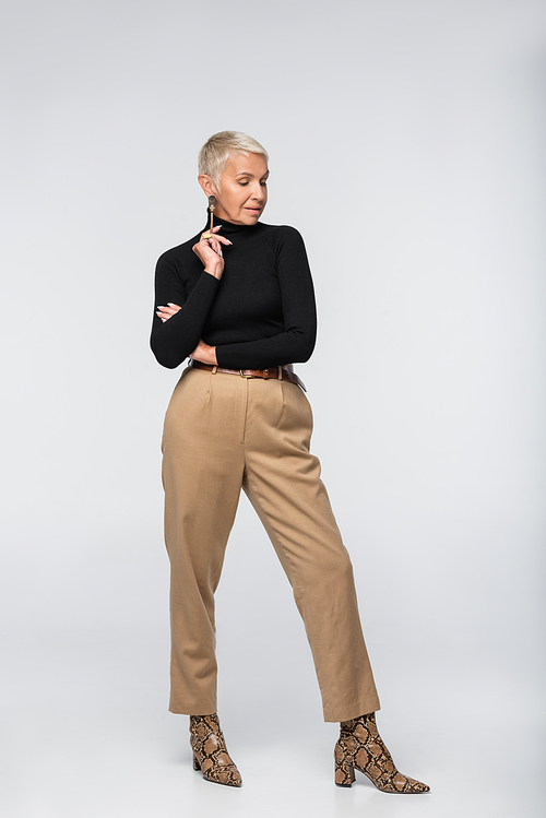 full length of pleased and elegant senior woman in beige pants, black turtleneck and animal  boots posing on grey