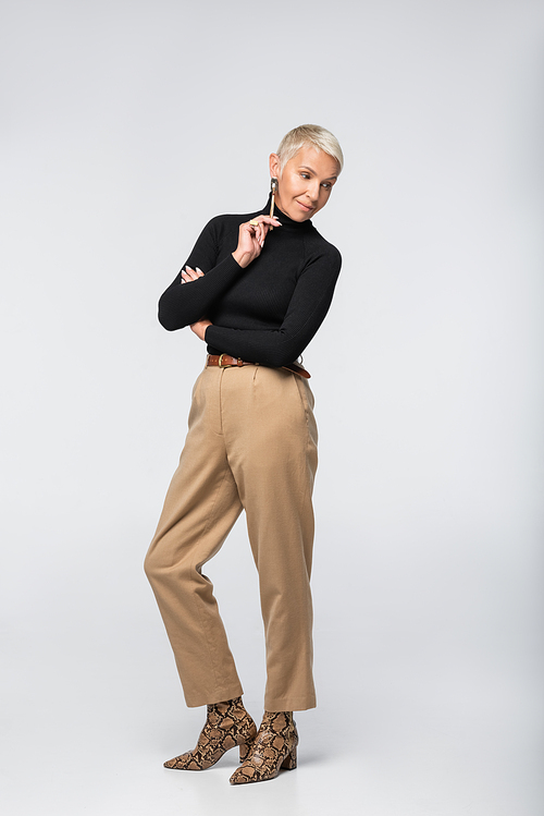 full length of pleased and stylish senior woman in beige pants and turtleneck posing on grey