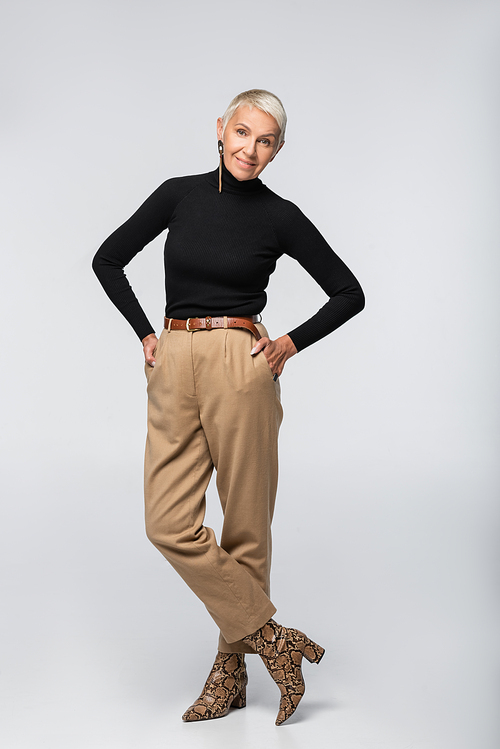 full length of smiling senior woman in beige pants, black turtleneck and animal  boots posing with hands in pockets on grey