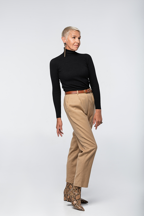 full length of stylish senior woman in beige pants, black turtleneck and animal  boots posing on grey