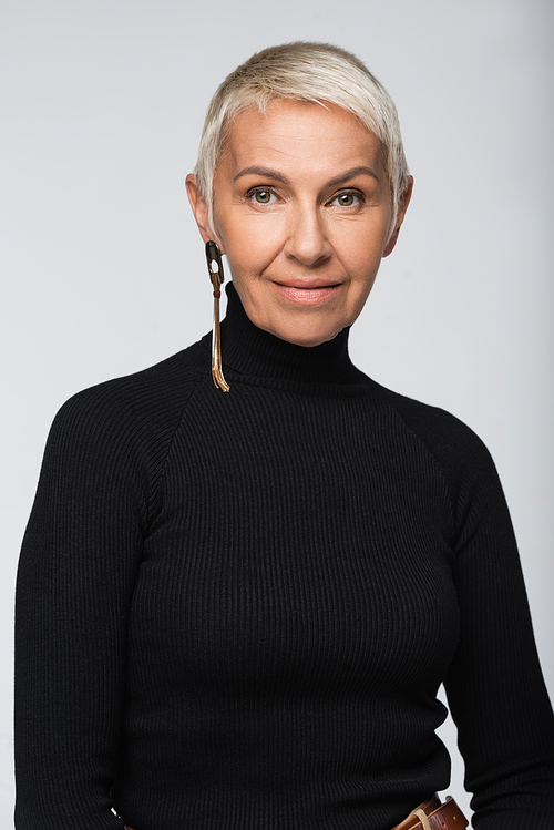 positive and senior woman with long earring and black turtleneck  isolated on grey