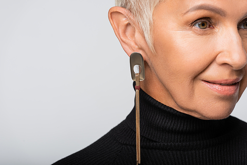 smiling and senior woman with long earring and black turtleneck looking away isolated on grey