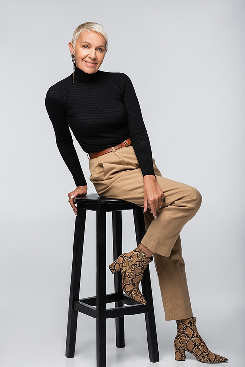 full length of cheerful senior woman in beige pants and turtleneck sitting on chair on grey