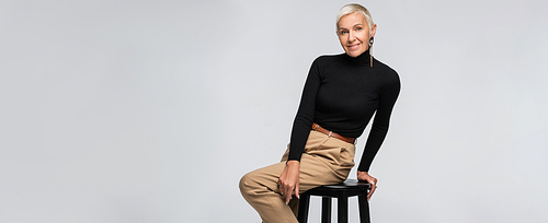 pleased and trendy senior woman in beige pants and turtleneck sitting on chair isolated on grey, banner