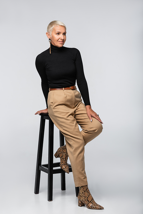 full length of smiling senior woman in beige pants and black turtleneck posing near chair on grey