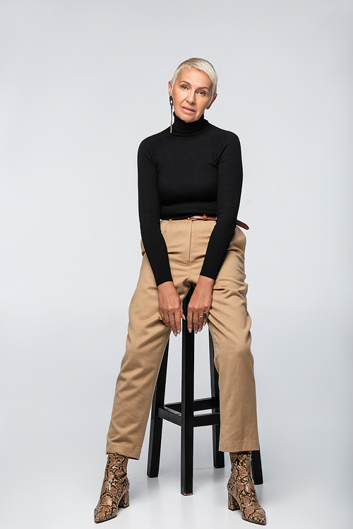full length of stylish senior woman in beige pants and turtleneck posing near chair on grey