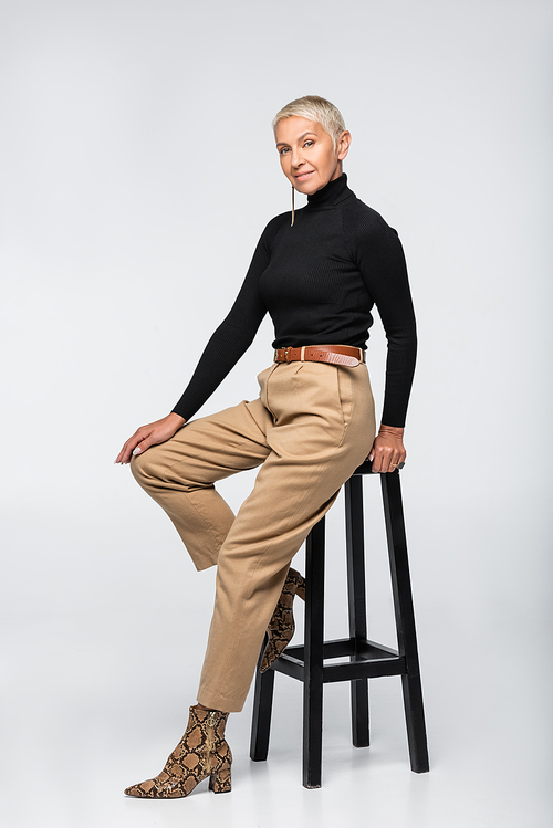 full length of pleased senior woman in beige pants and turtleneck posing near chair on grey