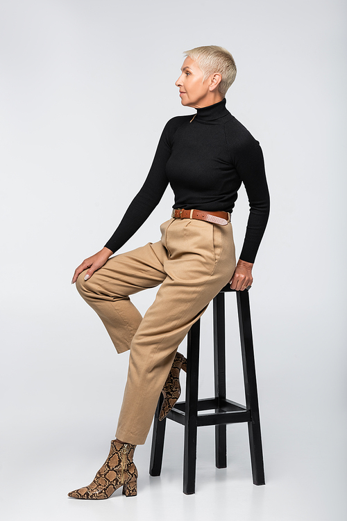 full length of trendy senior woman in beige pants and turtleneck posing near chair on grey